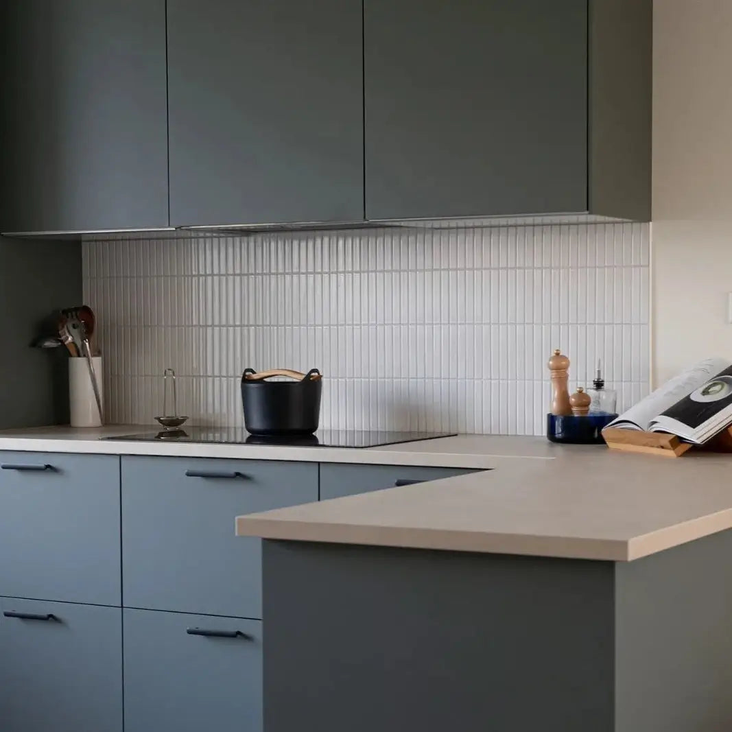 The Sevilla Mosaic Tiles, Finger KitKat design in a Glossy White Finish installed in a kitchen with dark cabinetry, under cabinet LED strip lighting, a white counter top slab with a cook top installed. Various kitchen utensils and a cookbook resting on the counter top.