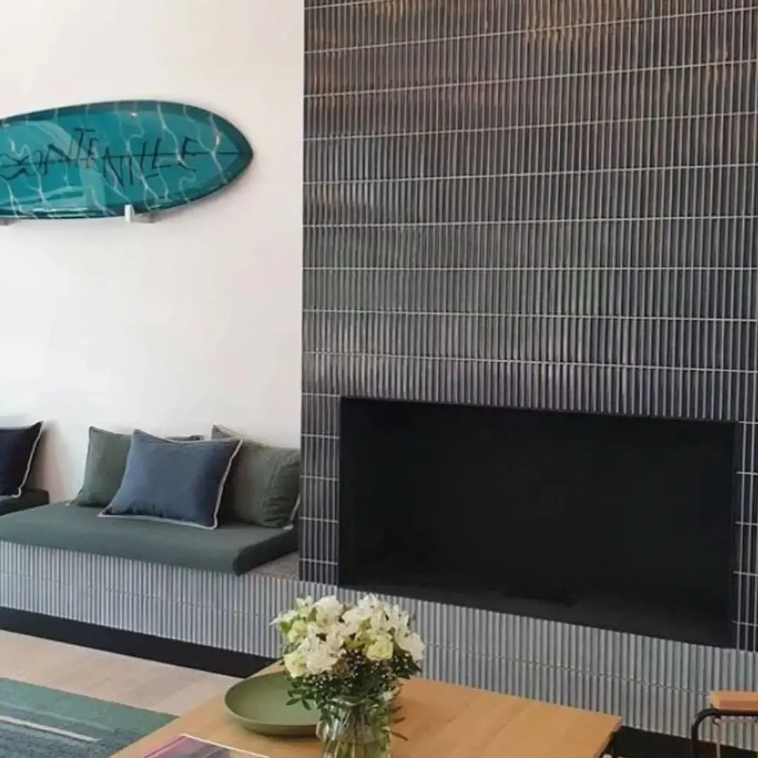 The Sevilla Mosaic Tiles, Original KitKat design in a Blue Gray Glossy, with a size of 20x145mm. Installed in a living/dining room setting with a with a television mounted on top, a table with a green plate, vase and flowers resting on it in front, a sitting area with green and blue cushions to the left and a surf board mounted on the wall above.