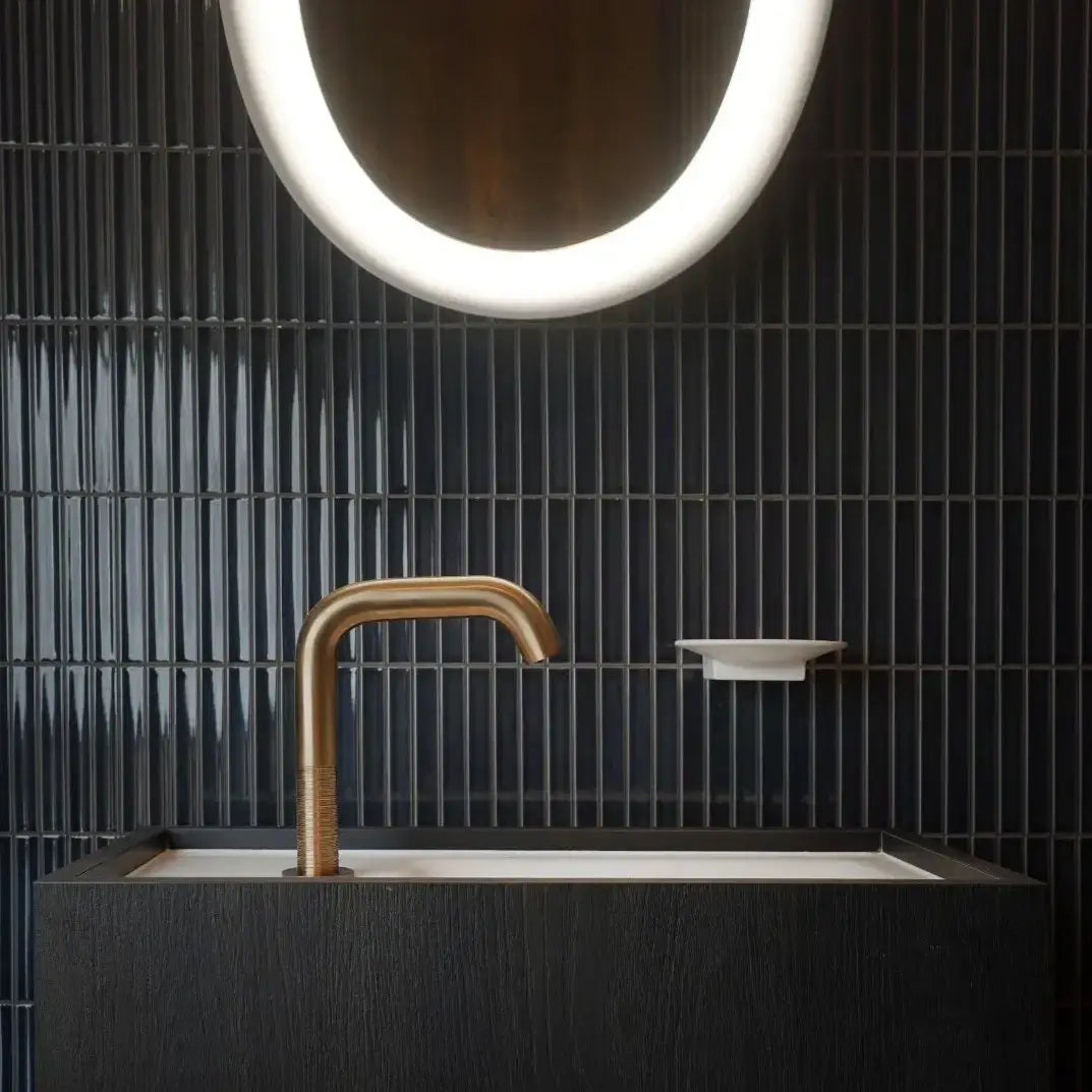 The Sevilla Mosaic Tiles, Original KitKat design in a Blue Gray Glossy, with a size of 20x145mm. Installed in a bathroom setting with a sink basin and bronze faucet in front and an LED mirror mounted above.