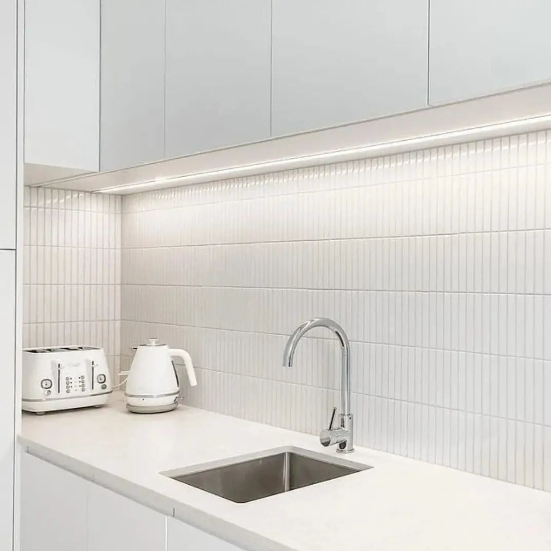 The Sevilla Mosaic Tiles, Finger KitKat design in a Glossy White Finish installed in a kitchen with white cabinetry, under cabinet LED strip lighting, a white counter top slab with a sink and chrome faucet installed. A white kettle and toaster appliance combo resting on the counter top.
