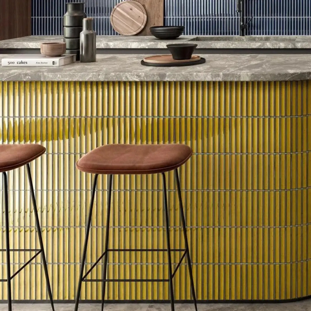 The Sevilla Mosaic Tiles, Original KitKat design in yellow, with a size of 22x145mm. Installed on a kitchen island bench with a stone counter top. Two stools standing in front of the bench. A Blue kit kat style mosaic tile in installed on the wall in the background.